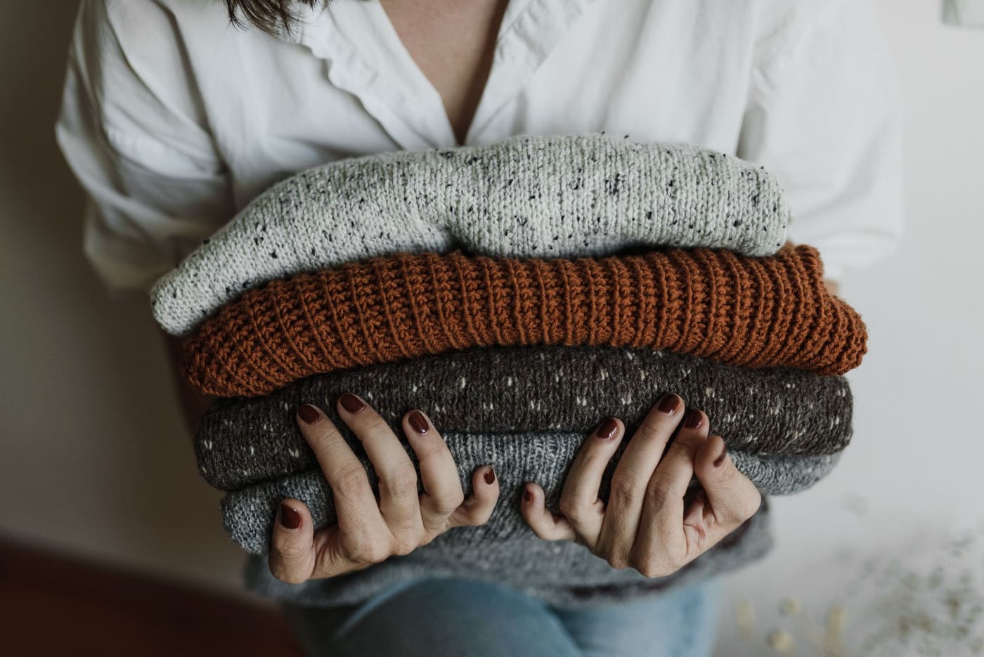 A person holding a stack of folded knit sweaters in grey, rust and brown