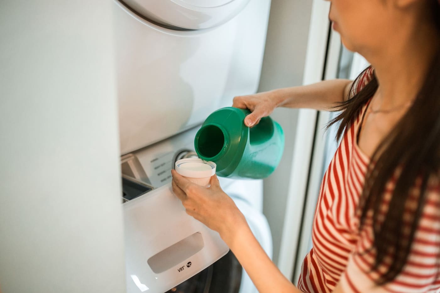 A woman pouring liquid detergent from a green bottle into its cap beside a washer